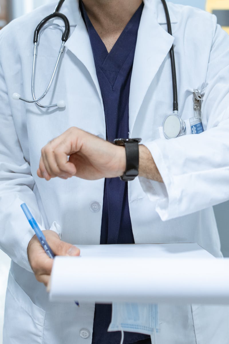 Doctor in lab coat checking wristwatch while holding medical chart in hospital corridor.